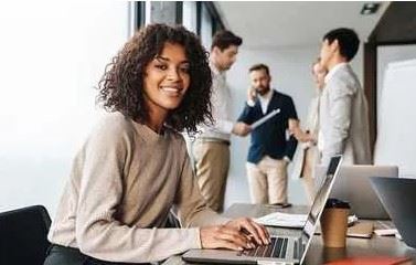 Lady at desk smiling with four coworkers in the background standing and chatting